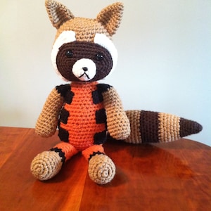 May include: A crocheted raccoon plush toy wearing an orange and black costume. The toy has a brown and tan striped tail.