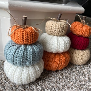 May include: A collection of handmade crochet pumpkins in various colors, including orange, white, blue, and beige. Each pumpkin has a small twig stem and twine detail, perfect for fall decor. The pumpkins are stacked in groups.
