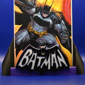 May include: A Batman comic book displayed in a black and purple stand. The comic features a dynamic illustration of Batman in a gray and black suit against a fiery background. The stand has a Batman logo.