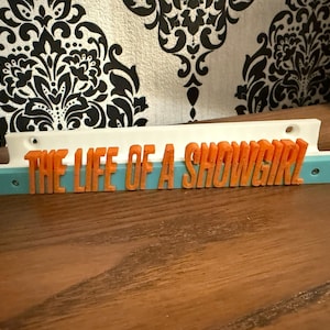 May include: A 3D-printed sign with the text "THE LIFE OF A SHOWGIRL" in orange letters. The sign has a white top, a light blue base, and is set against a patterned background.
