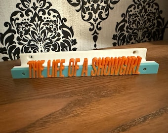 Könnte beinhalten: Ein 3D-gedrucktes Schild mit dem Text "THE LIFE OF A SHOWGIRL" in orangefarbenen Buchstaben. Das Schild hat eine weiße Oberseite, eine hellblaue Basis und befindet sich vor einem gemusterten Hintergrund.