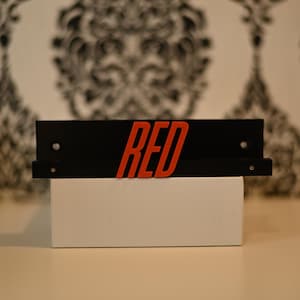 May include: A black shelf with the word "RED" in red, mounted on a white box. The shelf has two small holes and a lip to hold items. The background features a black and white patterned wallpaper.
