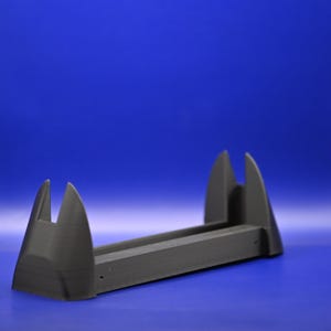 Op de afbeelding: Een zwarte plastic standaard met twee rechtopstaande punten, ontworpen om een klein object, zoals een telefoon of tablet, vast te houden.