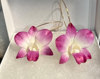 May include: A pair of gold hoop earrings with attached pink and white orchid flower pendants. The earrings are displayed in a white box with a textured, white interior.