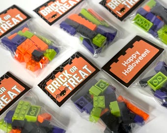 May include: Several clear plastic bags filled with colorful toy bricks. Each bag has a rectangular orange tag that reads "BRICK OR TREAT" with a spider and web graphic. One tag says "Happy Halloween!"