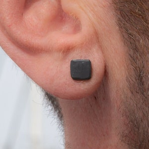 May include: A close-up of a person's ear with a small, square, black earring stud.
