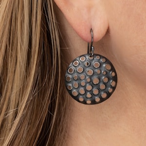 Peut inclure: Boucles d'oreilles en métal noir avec un design circulaire présentant un motif de petits trous.