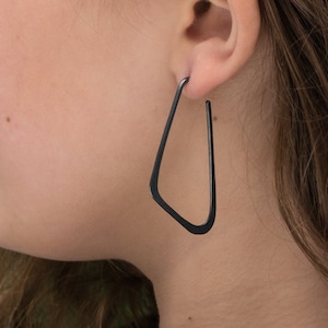 May include: A person wearing black triangle shaped hoop earrings. The earrings are made of metal and have a matte finish.