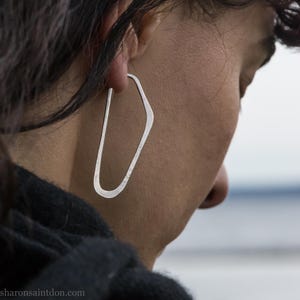 May include: A close-up of a silver earring with an irregular geometric shape. The earring is a single piece of metal with a hammered texture. The earring is worn on the ear of a person, with the background blurred.