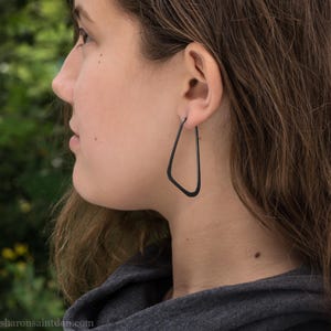 May include: A side profile view of a person wearing a pair of dark gray, geometric hoop earrings. The earrings have a unique, angular shape, with a straight top and a slanted bottom. The background is blurred, with green foliage visible.