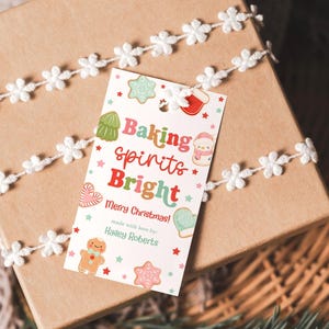 May include: A wrapped gift box with a festive Christmas tag. The tag reads "Baking spirits Bright" with colorful illustrations of gingerbread men, cookies, and a snowman. The box is tied with a white floral ribbon.