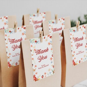 May include: Brown paper gift bags with floral "Thank You" tags. The tags feature red text, butterflies, ladybugs, and flowers. Each tag is clipped to the bag with a small wooden clothespin. The text "Love, Kate & Daniel" is also on the tags.