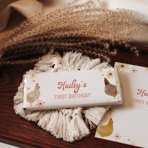 May include: A chocolate bar wrapper with the text "Hailey's FIRST BIRTHDAY". The wrapper features illustrations of chickens and flowers. The bar rests on a macrame coaster, with dried plants in the background. The overall theme is a birthday.
