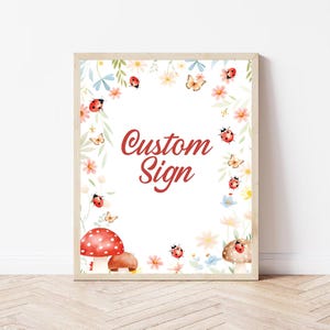 May include: A framed print with a floral and insect design. The print features a white background with a border of flowers, butterflies, and ladybugs. The words "Custom Sign" are written in red script.