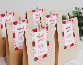 Puede incluir: Bolsas de papel marrón con etiquetas de agradecimiento rosas y blancas con un patrón de fresas y flores. Las etiquetas dicen "¡Gracias!" y "Por celebrar el primer cumpleaños de Scarlett con nosotros. Con cariño, la familia Williams."