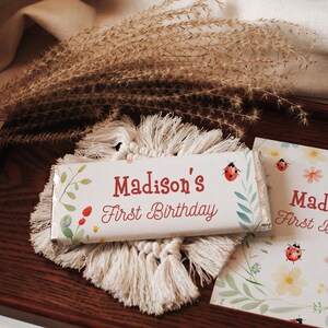 May include: A white chocolate bar wrapper with the text "Madison's First Birthday" in red script, decorated with floral and ladybug illustrations. The bar rests on a macramé coaster, with a matching card nearby.