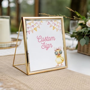 May include: A gold metal framed sign with the words "Custom Sign" in pink script. The sign features a watercolor illustration of a yellow duckling wearing sunglasses and a pink bow. The background includes a garland of pink and yellow pennants.
