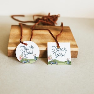 May include: Two gift tags with brown string. One is round, the other square. Both have the text "Thank You!" in script. The tags feature a golf cart, balloons, and a golf course scene. The tags are on a wooden surface.