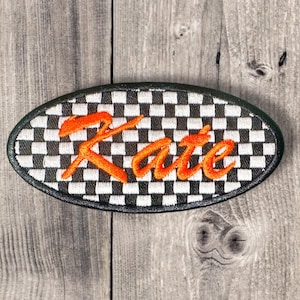 Op de afbeelding: Ovale patch met een zwart-wit geblokt patroon en de naam "Kate" geborduurd in oranje draad. De patch heeft een zwarte rand en is geplaatst tegen een verweerde houten achtergrond.