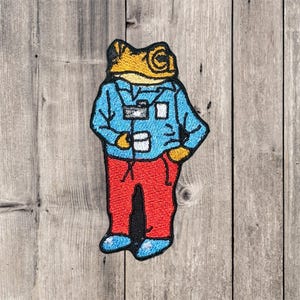 Pode incluir: Patch bordado com um desenho de um sapo de desenho animado usando uma camisa azul, calças vermelhas e sapatos azuis. O sapo tem uma cabeça amarela e segura um pequeno objeto branco. Em fundo de madeira.