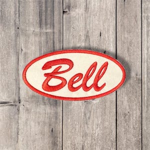 May include: An oval-shaped patch with the word "Bell" embroidered in red script. The patch has a cream-colored background and a red border. The patch is set against a weathered, gray wooden background.