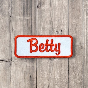May include: A white rectangular patch with a red border. The word "Betty" is embroidered in red on the patch.