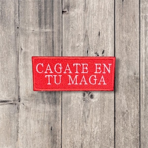 May include: A rectangular red patch with white text that reads "CAGATE EN TU MAGA". The patch is set against a weathered, gray wooden background. The text is centered and in a sans-serif font.