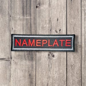 Könnte beinhalten: Ein rechteckiger Aufnäher mit dem Wort "NAMEPLATE" in Rot auf schwarzem Hintergrund gestickt. Der Aufnäher hat einen weißen Rand und einen schwarzen Rand, vor einem Holzgrund.