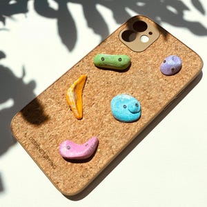 Puede incluir: Una funda de teléfono con textura de corcho, adornada con adornos coloridos en forma de roca pintada. La funda presenta un recorte de cámara marrón y el texto "DESIGNER ZIYING". Las rocas son en tonos de verde, amarillo, azul, morado y rosa.