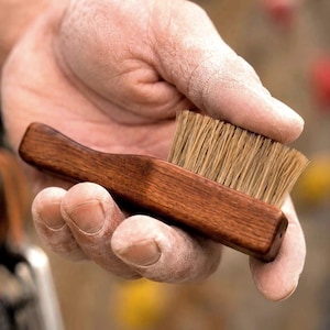 May include: A wooden-handled brush with light brown bristles. The brush is held in a hand dusted with a white powder. The wood handle is a dark brown colour.