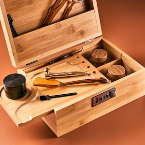 May include: A bamboo stash box with a combination lock, open to reveal various tools and containers. Includes a black grinder, a small brush, a gold-colored pipe, and two glass jars with wooden lids. The box is on a brown surface.