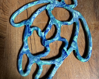 May include: A ceramic trivet featuring an abstract, organic, open-work design with a glossy finish. The surface displays a marbled pattern of blended deep blue, light blue, and bright green colours, creating a fluid, interconnected network of loops.