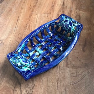 May include: A blue and green ceramic bread basket with a woven design. The basket is shaped like a boat and has a decorative handle.