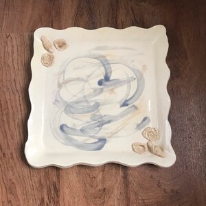 May include: Square ceramic serving platter with a scalloped edge. The off-white platter features blue and tan brushstrokes and is decorated with small seashell accents. A decorative piece for serving food.