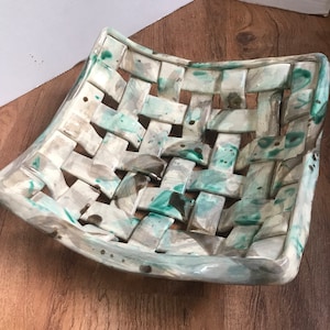May include: A woven ceramic bowl with a light gray and green glaze. The bowl has a square shape with rounded edges and a woven pattern.
