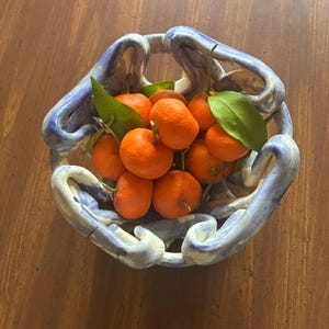 May include: A ceramic bowl with a blue and white design holds a collection of bright orange tangerines with green leaves. The bowl has a unique, open design with curved handles. The tangerines are fresh and vibrant, creating a colorful display.
