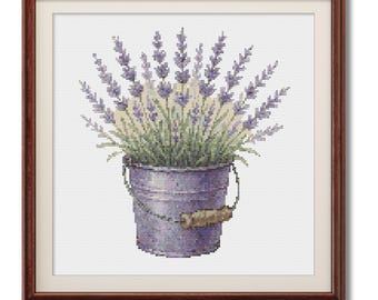May include: Cross-stitch artwork of lavender in a galvanized bucket with a wooden handle, set in a brown framed print. The lavender has purple flower spikes and green foliage. The bucket is a muted purple color.