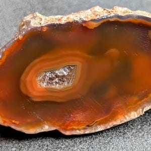 May include: A polished agate stone slice with concentric bands of orange, brown, and white. The center features a crystalline cavity. The stone has a rough, natural edge and is displayed on a gray surface.