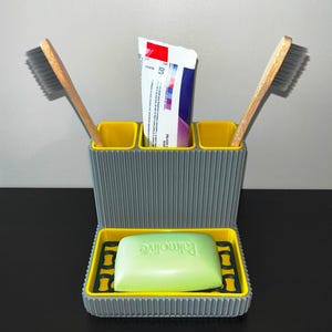 May include: Gray and yellow bathroom organizer with a ribbed texture. It has three slots for toothbrushes and a compartment for toothpaste. A bar of soap is in the bottom tray.