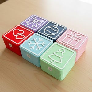 May include: A collection of six colorful square boxes with white embossed designs. The designs include a bow, ornament, gift, hearts, snowflake, and Christmas tree. The boxes are arranged on a light wooden surface.