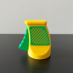 May include: A yellow and green plastic toy with a handle and a textured green piece on the side. The toy has a rectangular green section with diagonal lines and circular details. The toy is on a black surface.