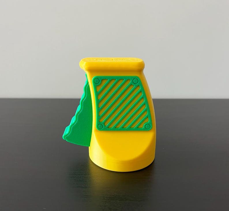 May include: A yellow and green plastic toy with a handle and a textured green piece on the side. The toy has a rectangular green section with diagonal lines and circular details. The toy is on a black surface.