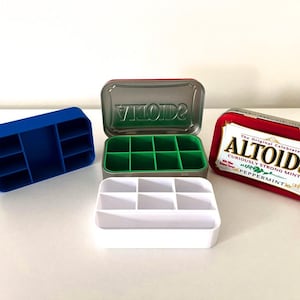 May include: A collection of rectangular storage inserts for Altoids tins, including a blue eight-compartment tray, a green eight-compartment tray inside a metal tin, a white six-compartment tray, and a closed red Altoids Peppermint mint tin.
