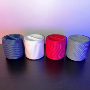 May include: Four small, cylindrical containers with different textured surfaces. The containers are blue, white, red, and gray.