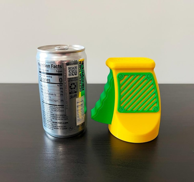 May include: A silver soda can next to a yellow and green can opener. The can opener has a yellow body with green accents and a green handle. The can has a nutrition facts label.