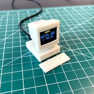 May include: A white miniature desktop computer with a blue screen displaying "AAPL $235.00 +$2.85". The computer is on a green cutting mat with a white keyboard.