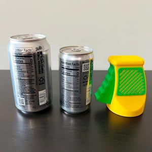 May include: Two silver soda cans and a yellow and green can opener sit on a dark surface. The cans have nutrition facts labels. The can opener has a green ramp and a yellow body with a green grid design.