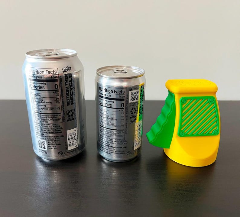 May include: Two silver soda cans and a yellow and green can opener sit on a dark surface. The cans have nutrition facts labels. The can opener has a green ramp and a yellow body with a green grid design.