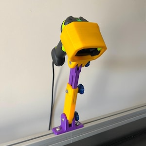 Puede incluir: Un soporte para escáner impreso en 3D de color amarillo y morado, fijado a una vía de plata. El escáner es negro con una cubierta protectora amarilla y un botón verde.