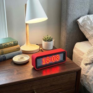 May include: A red digital alarm clock displaying 6:08 PM sits on a wooden nightstand. A white lamp with a wooden base and a small succulent in a white pot are also on the nightstand. A stack of books and a small white dish are also visible.